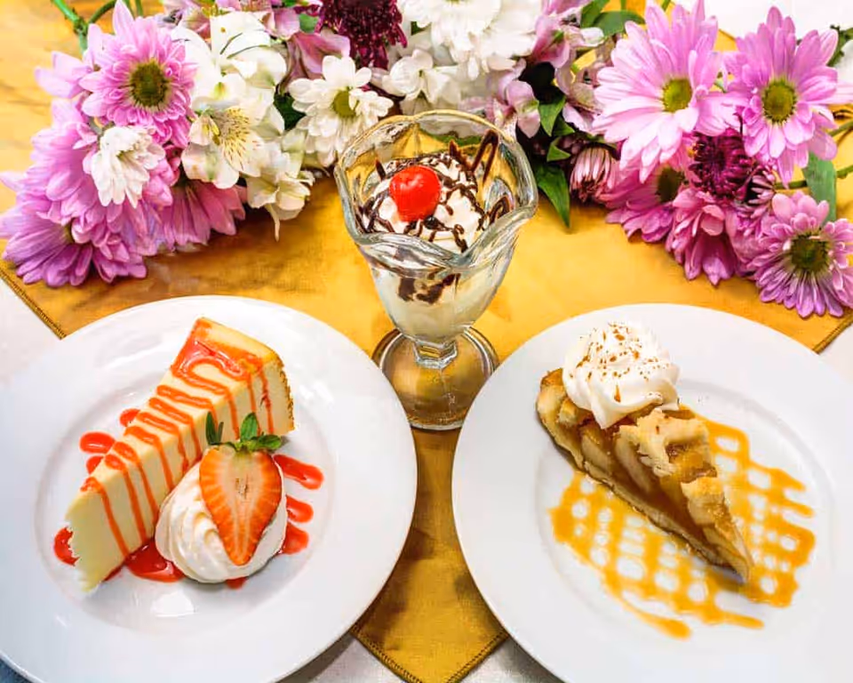 Two dessert plates and a sundae glass on a yellow tablecloth with a bouquet of pink and white flowers in the background. One plate has a slice of cheesecake with strawberry sauce, whipped cream, and a strawberry half. The other plate has a slice of apple pie with caramel drizzle and whipped cream. The sundae glass contains vanilla ice cream topped with chocolate syrup and a cherry.