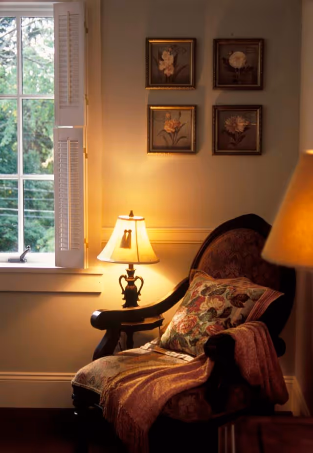 Upholstered armchair with a floral pillow and throw beside a lit table lamp under framed floral pictures next to a window.