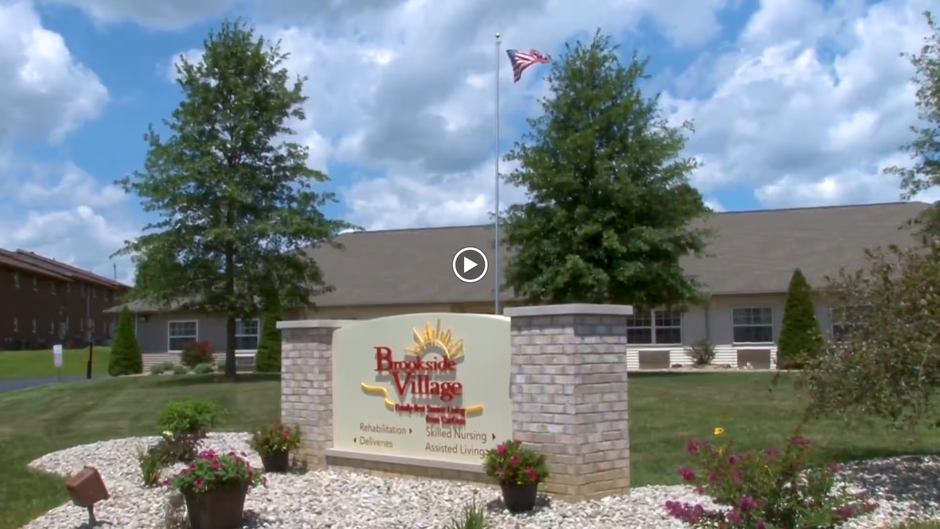 Landscaped front lawn with a stone sign for Brookside Village Family-first Senior Living, the facility building and an American flag in the background.