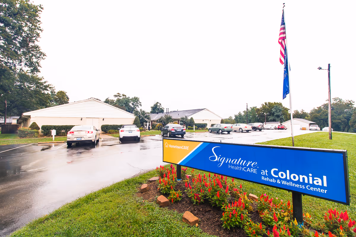 Exterior view of Signature HealthCARE at Colonial Rehab & Wellness Center with a parking lot, several cars, a flagpole with American and state flags, and a blue sign with the facility name surrounded by red flowers and green grass.