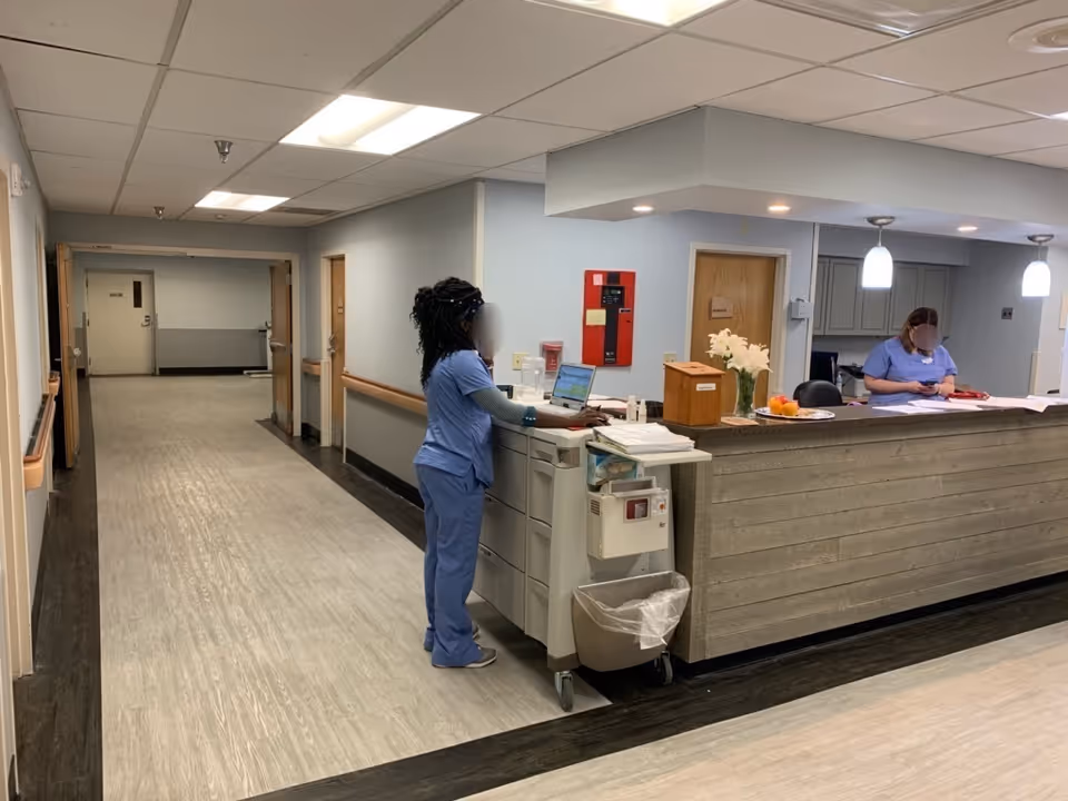 Nurse station in a facility hallway with two staff in scrubs working at a reception desk.
