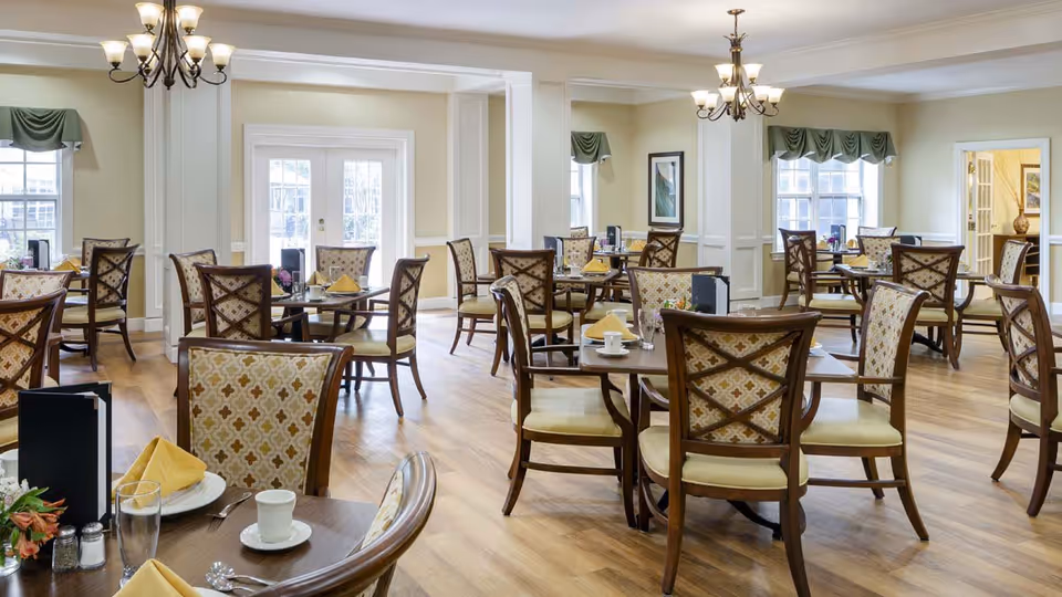 A bright and spacious dining room with multiple wooden tables and cushioned chairs arranged neatly. Each table is set with plates, cups, glasses, silverware, and yellow folded napkins. The room features light-colored walls, large windows with green valances, and two chandeliers hanging from the ceiling. The floor is wooden, and there are decorative elements such as framed artwork on the walls.