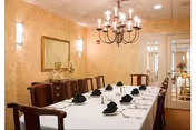 A formal dining room with a long rectangular table covered with a white tablecloth, set with black folded napkins and white plates. The room has beige patterned wallpaper, a chandelier hanging from the ceiling, a large mirror on one wall, and wooden chairs surrounding the table.
