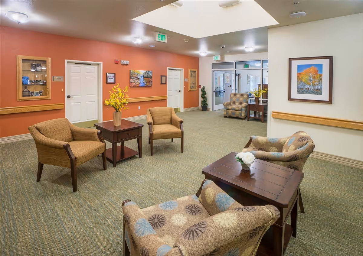 A well-lit seating area in a senior living facility with several armchairs arranged around wooden tables. The walls are painted in warm tones with artwork and a display case. There is a carpeted floor and ceiling lights, with glass doors leading to another area in the background.