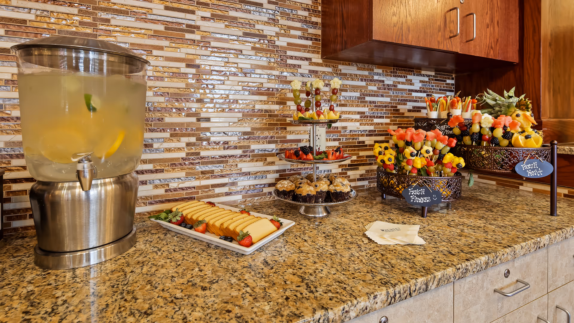 A countertop with a large beverage dispenser filled with lemon water, a rectangular plate with sliced pound cake and strawberries, a tiered tray with muffins and fruit skewers, and two decorative baskets filled with assorted fresh fruit skewers labeled 'Fresh Flowers' and 'Fresh Fruit'. The background features a mosaic tile backsplash and wooden cabinets above.