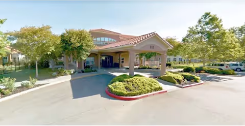 Front entrance of a senior living community with a covered porte-cochere, driveway, and landscaped trees and shrubs.
