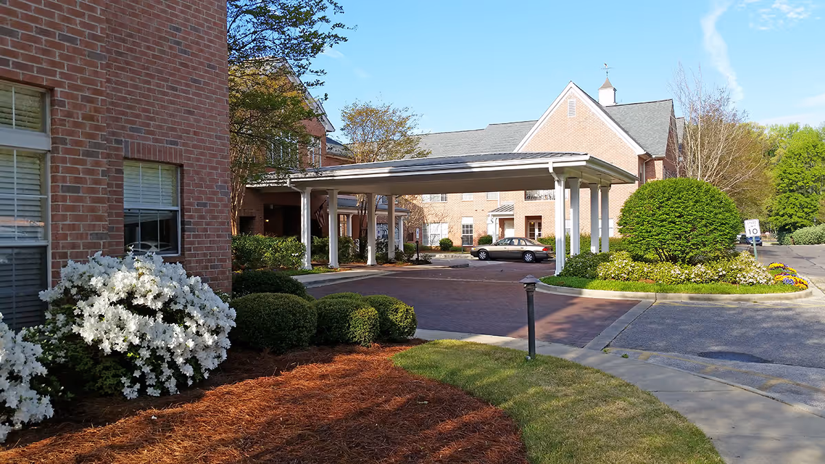 Exterior view of a senior living facility with brick buildings, a covered driveway entrance, neatly trimmed bushes, white flowering plants, and a clear blue sky.