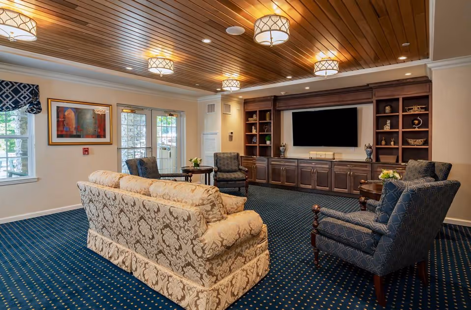 A cozy living room with a patterned beige sofa and two blue armchairs arranged around a small round table. The room features a wooden ceiling with multiple ceiling lights, a large built-in wooden entertainment center with a flat-screen TV, and decorative shelves. There are windows and glass doors letting in natural light, and the floor is covered with a blue carpet with a small dotted pattern.