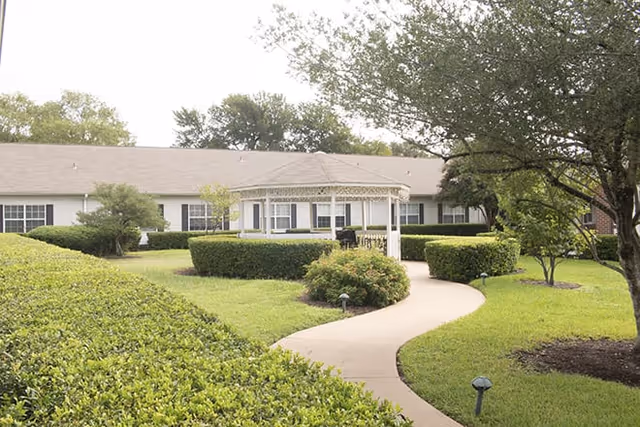 A well-maintained garden area with a curved concrete pathway leading to a white gazebo surrounded by neatly trimmed bushes and trees, in front of a single-story building with multiple windows.