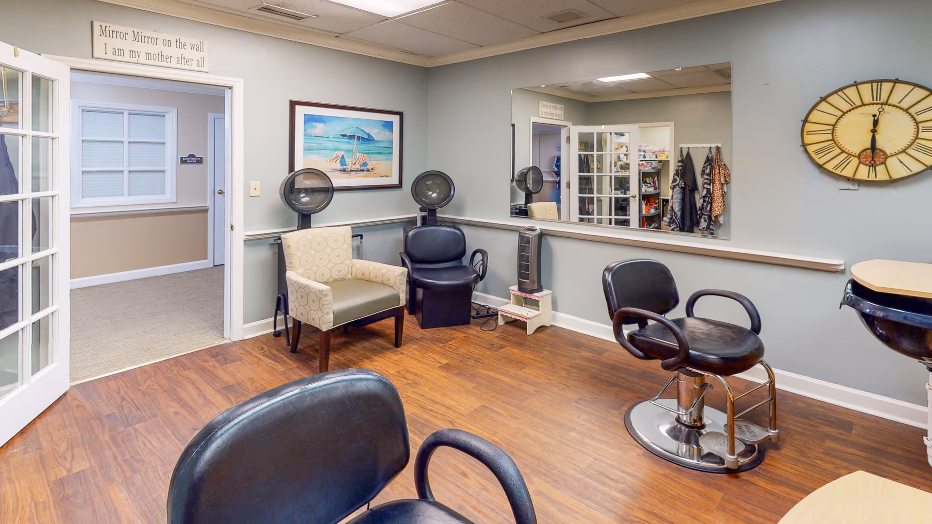 A salon-style room with styling chairs, hooded hair dryers, a large mirror, seating, and a wall clock.