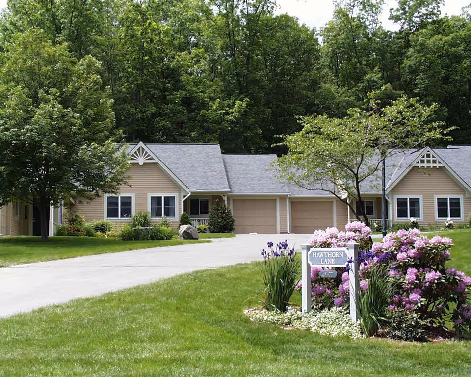 A peaceful residential street named Hawthorn Lane in Lathrop Retirement Community, featuring beige single-story homes with attached garages, surrounded by green lawns, trees, and blooming purple and pink flowers.