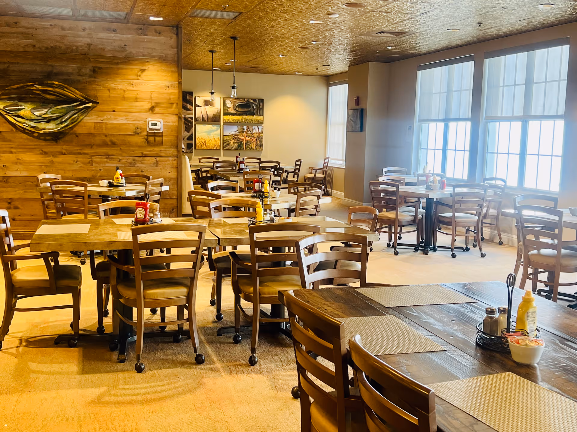 Bright dining room with multiple wooden tables and chairs, placemats and condiments, large windows, and a wood-paneled accent wall.