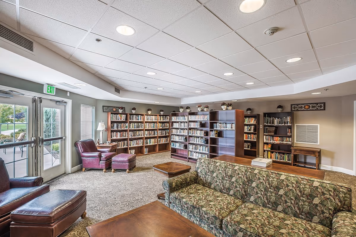 A comfortable library lounge with bookshelves, upholstered chairs and sofas, tables, and glass doors leading outside.