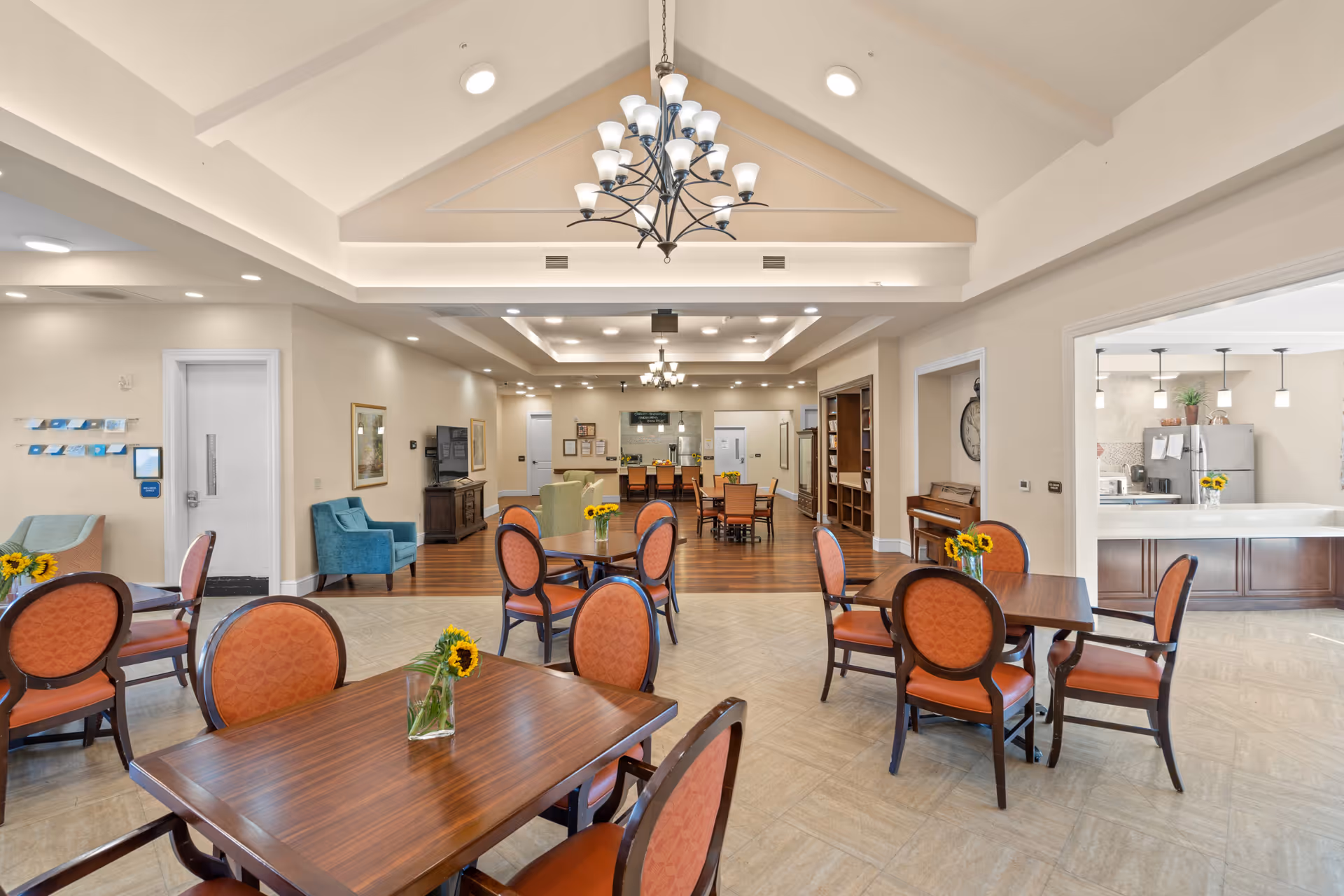 Bright dining and common area with multiple wooden tables and upholstered chairs, a chandelier overhead, and an open kitchen in the background.