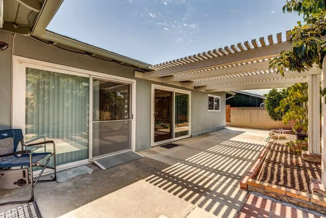 Concrete patio with a pergola casting striped shadows, sliding glass doors, a grill and raised planter beds at the rear of a single-story house.