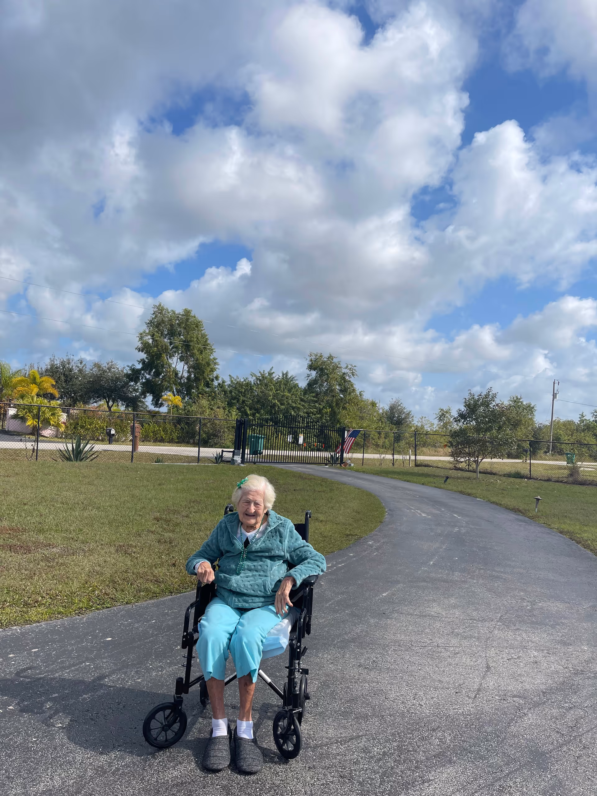 An elderly woman sitting in a wheelchair on a paved path outdoors, surrounded by grass and trees under a partly cloudy blue sky.
