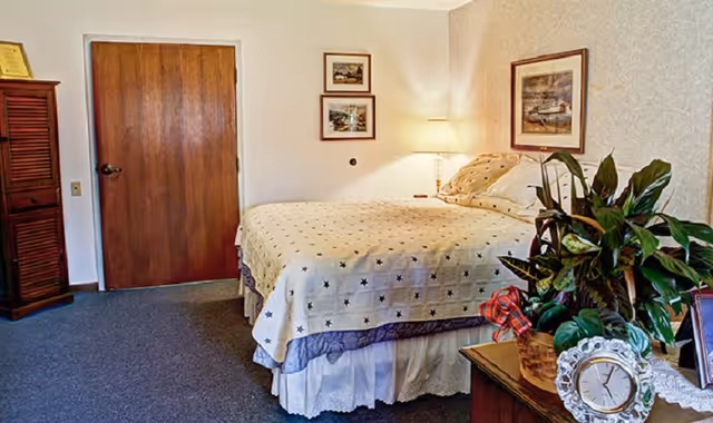 A cozy bedroom with a bed covered in a cream-colored quilt with star patterns. There is a wooden door on the left side, a wooden cabinet, and a nightstand with a lamp, a clock, a framed photo, and a potted plant. Two framed pictures hang on the wall above the bed.