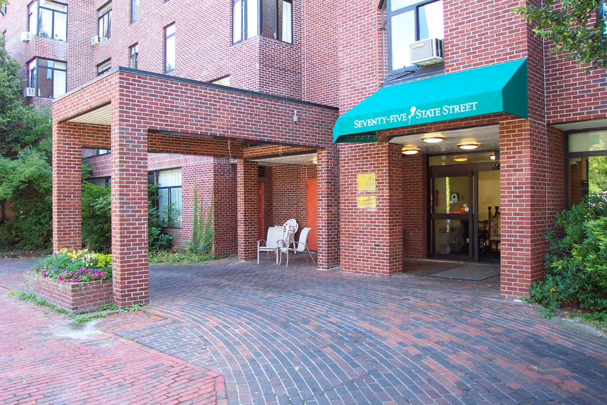 Entrance of a brick building with a green awning that reads 'Seventy-Five State Street'. There is a covered driveway area with two white chairs and a small table. The entrance has glass doors and is surrounded by greenery and flower beds.
