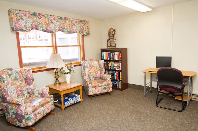 A cozy room with two floral-patterned armchairs, a wooden side table with a lamp and a vase of flowers, a bookshelf filled with books, and a small desk with a computer and a chair. The room has beige walls, a window with a floral valance, and carpeted flooring.