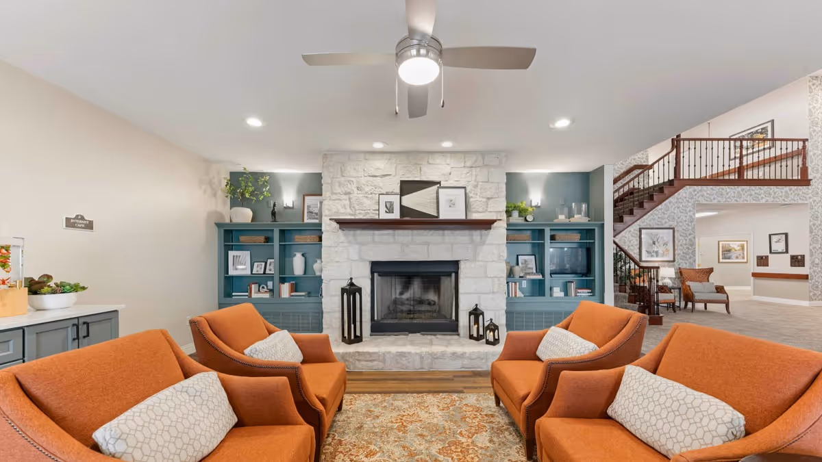 A cozy living room area with four orange armchairs arranged around a patterned rug facing a stone fireplace. The fireplace has a wooden mantel with framed artwork and decorative lanterns on the hearth. Built-in blue shelves flank the fireplace, holding books, vases, and plants. A ceiling fan with a light is above, and a staircase with wooden railing is visible in the background.