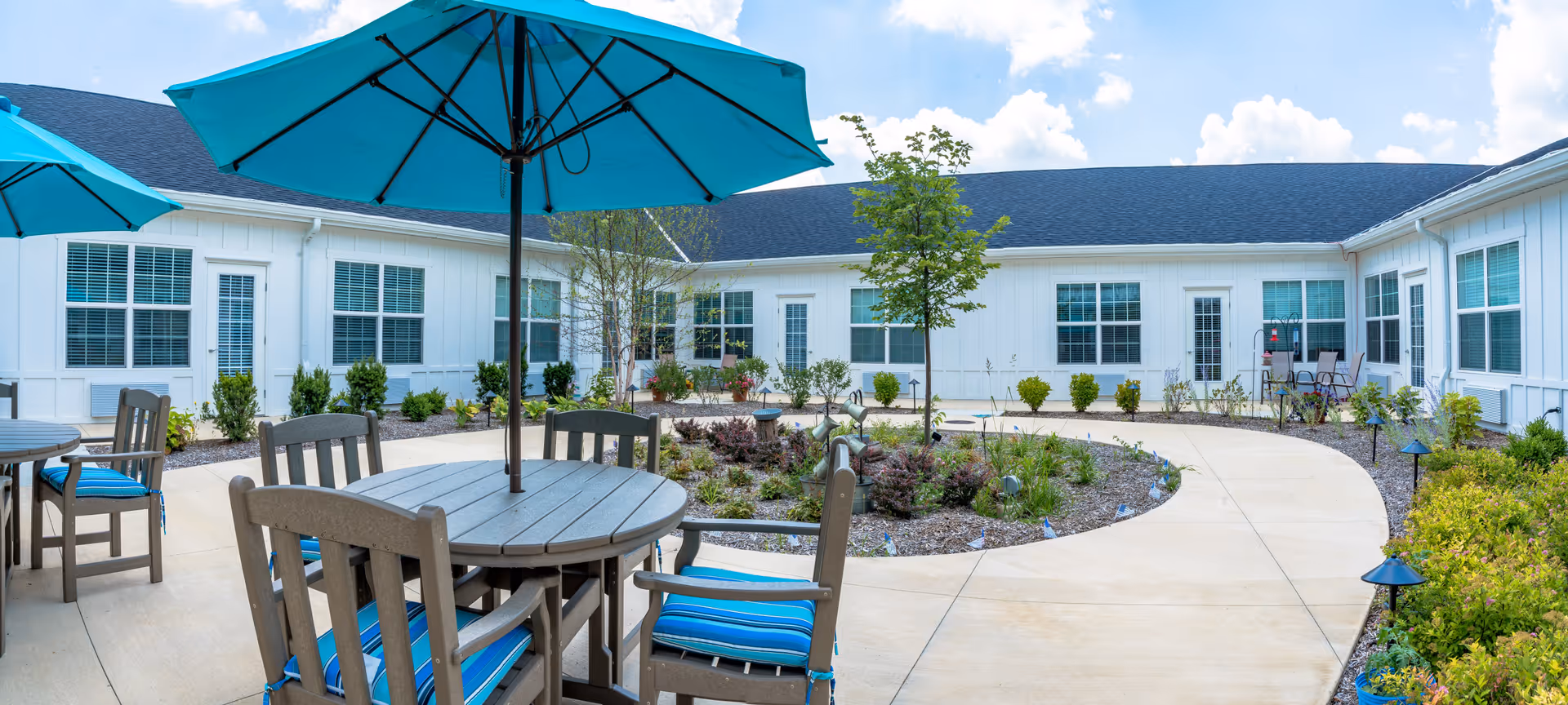 Outdoor courtyard area at Sugar Fork Crossing featuring round tables with blue umbrellas and matching blue cushioned chairs, surrounded by a curved walkway and landscaped garden beds with small trees and shrubs, adjacent to a white building with multiple windows and doors.