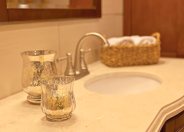 Bathroom vanity countertop with sink and faucet, two decorative glass votives, and a wicker basket holding towels.