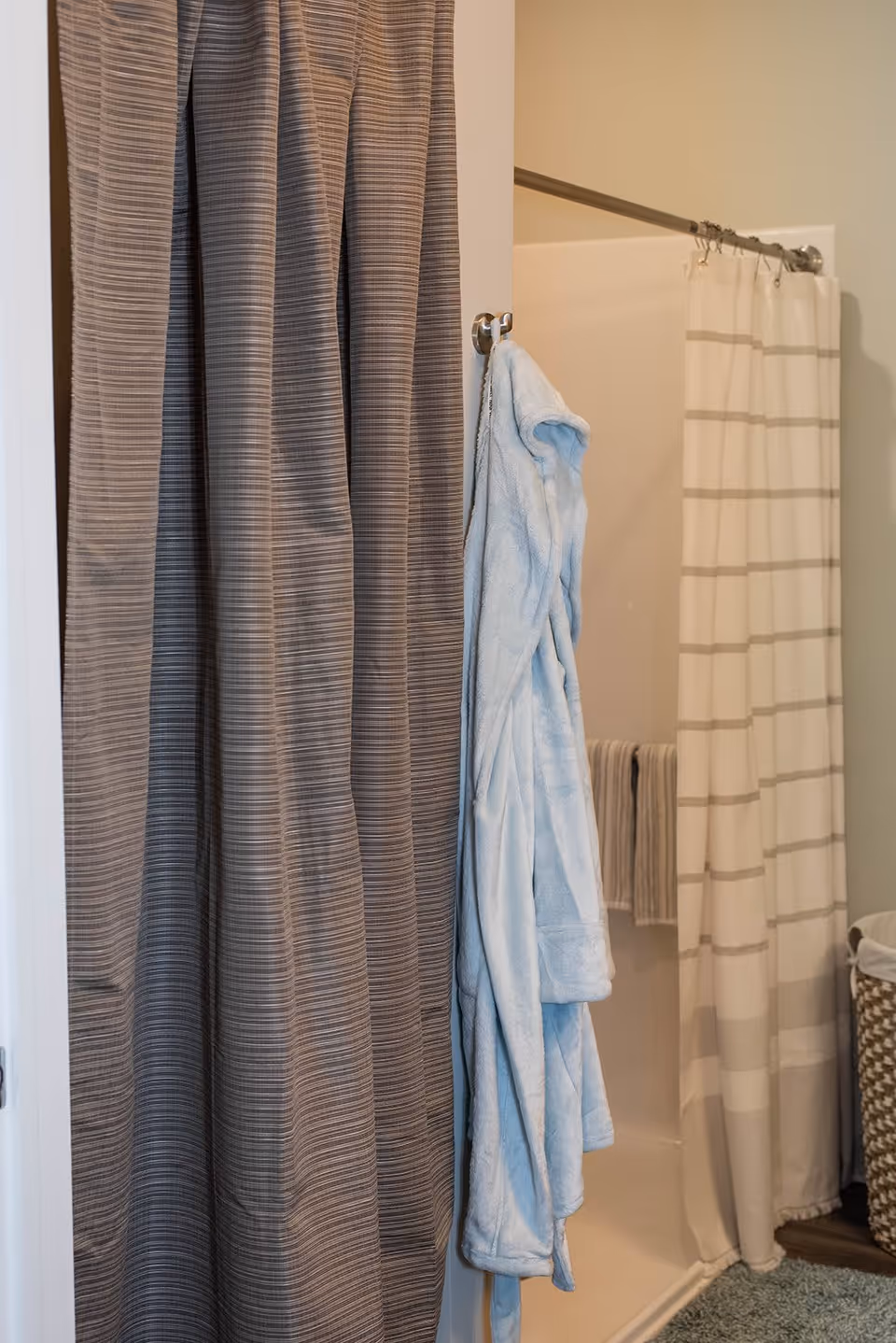 Shower area with striped shower curtains, a light blue bathrobe hanging on a hook, and a laundry basket nearby.