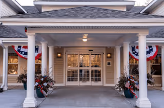 Entrance of All American Assisted Living at Kingston featuring a covered porch with white columns, patriotic red, white, and blue bunting decorations, and planters with American flags on either side of the double glass doors.
