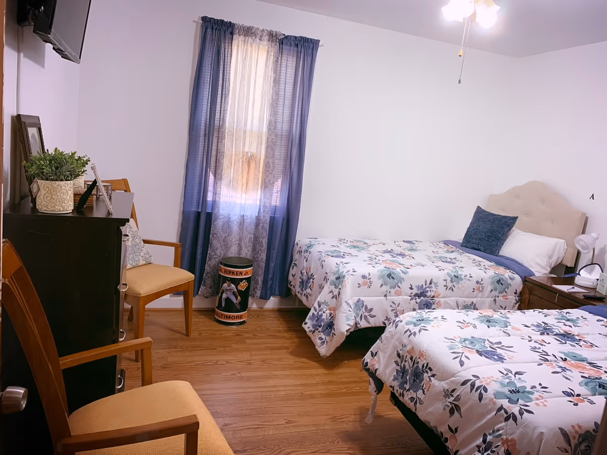 Bright bedroom with two twin beds with floral bedding, a dresser and chairs, and a window with blue curtains.