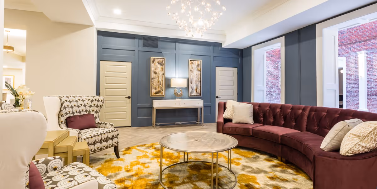 Bright communal living room with a curved burgundy sofa, patterned armchairs, round coffee table, and decorative wall with doors and artwork.