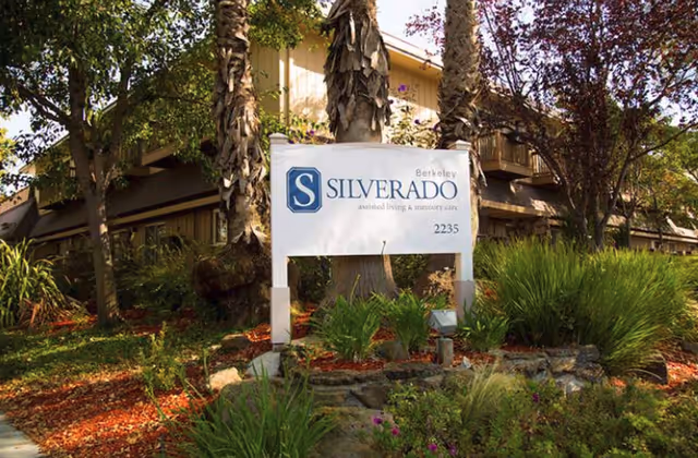 Outdoor view of Silverado Berkeley Memory Care Community sign surrounded by trees, plants, and landscaping with a building partially visible in the background.