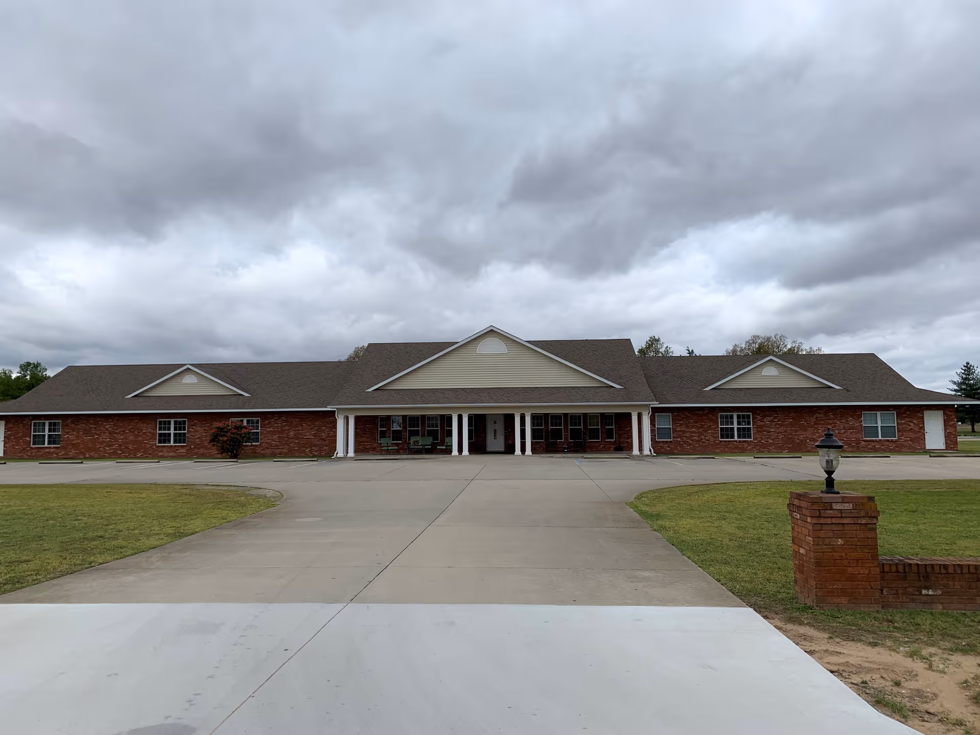 Front view of a single-story brick assisted living building with a covered entrance and wide driveway under a cloudy sky.