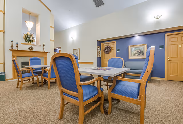 A cozy common area with a table and four blue cushioned wooden chairs in the foreground. In the background, there is a fireplace with decorative items and a floral arrangement above it. The walls are painted in light colors with some artwork and a wreath on a door. The room has carpeted flooring and soft lighting fixtures on the walls.