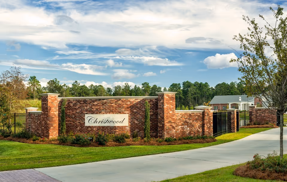 Brick entrance wall with a 'Christwood' sign at the driveway entrance to a retirement community, with landscaping and buildings visible behind it.