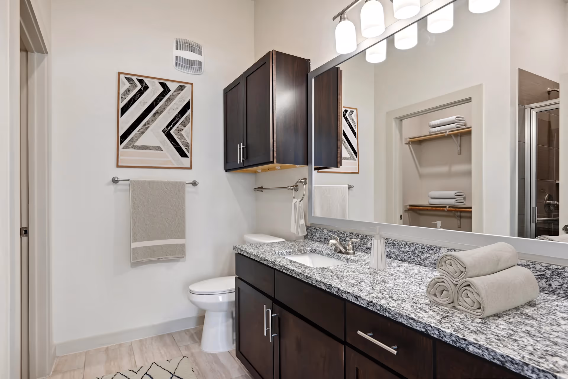 Modern bathroom with granite countertop, dark wood cabinets, a toilet, large mirror, and neatly rolled towels.