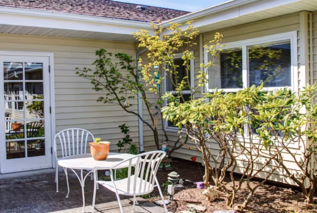 Outdoor patio area with a white metal table and two matching chairs. A small potted plant is on the table. The patio is adjacent to a beige building with a window and a glass door. There are green shrubs and small trees planted along the building's exterior wall.
