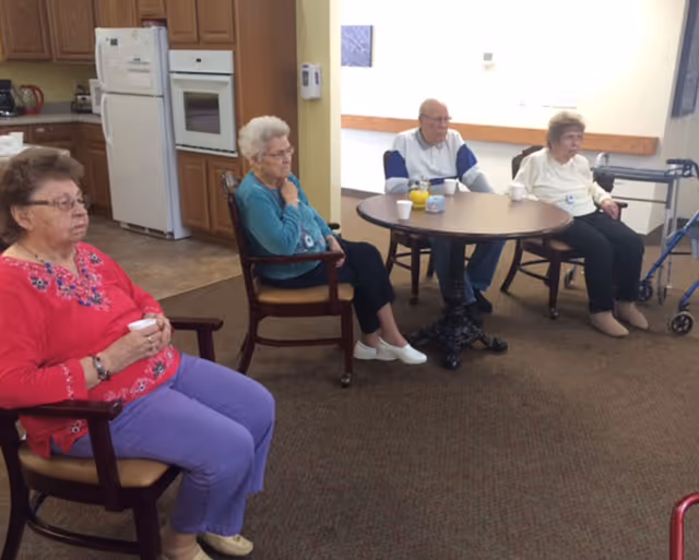 Four older adults sit in chairs around a small round table in a senior living facility common area with a kitchenette in the background.