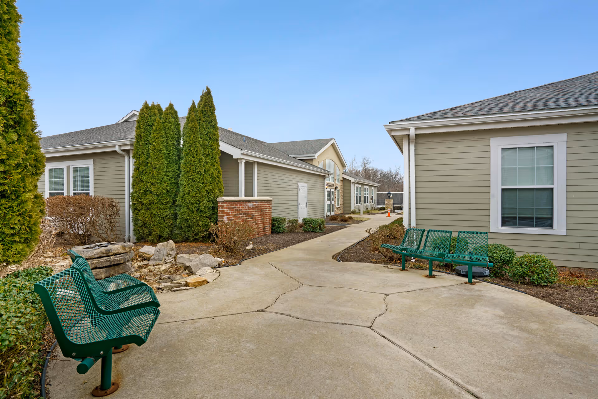 Outdoor walkway area between single-story buildings with green metal benches on either side, landscaped with bushes, rocks, and tall evergreen trees under a clear blue sky.
