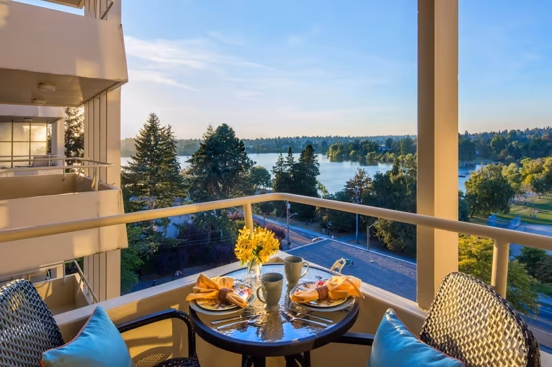 A balcony with a small round table set for two with plates, napkins, cups, and a vase of yellow flowers. Two wicker chairs with blue cushions are positioned around the table. The balcony overlooks a scenic view of a lake, trees, and a park under a clear sky.
