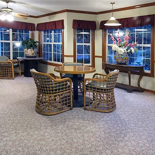 A cozy interior common area with a round wooden table surrounded by four wicker chairs with cushions. The room has large windows with maroon valances, beige walls with wooden trim, and carpeted flooring. There are two side tables, one with a large potted plant and the other with a decorative floral arrangement. Ceiling lights and a ceiling fan provide illumination.