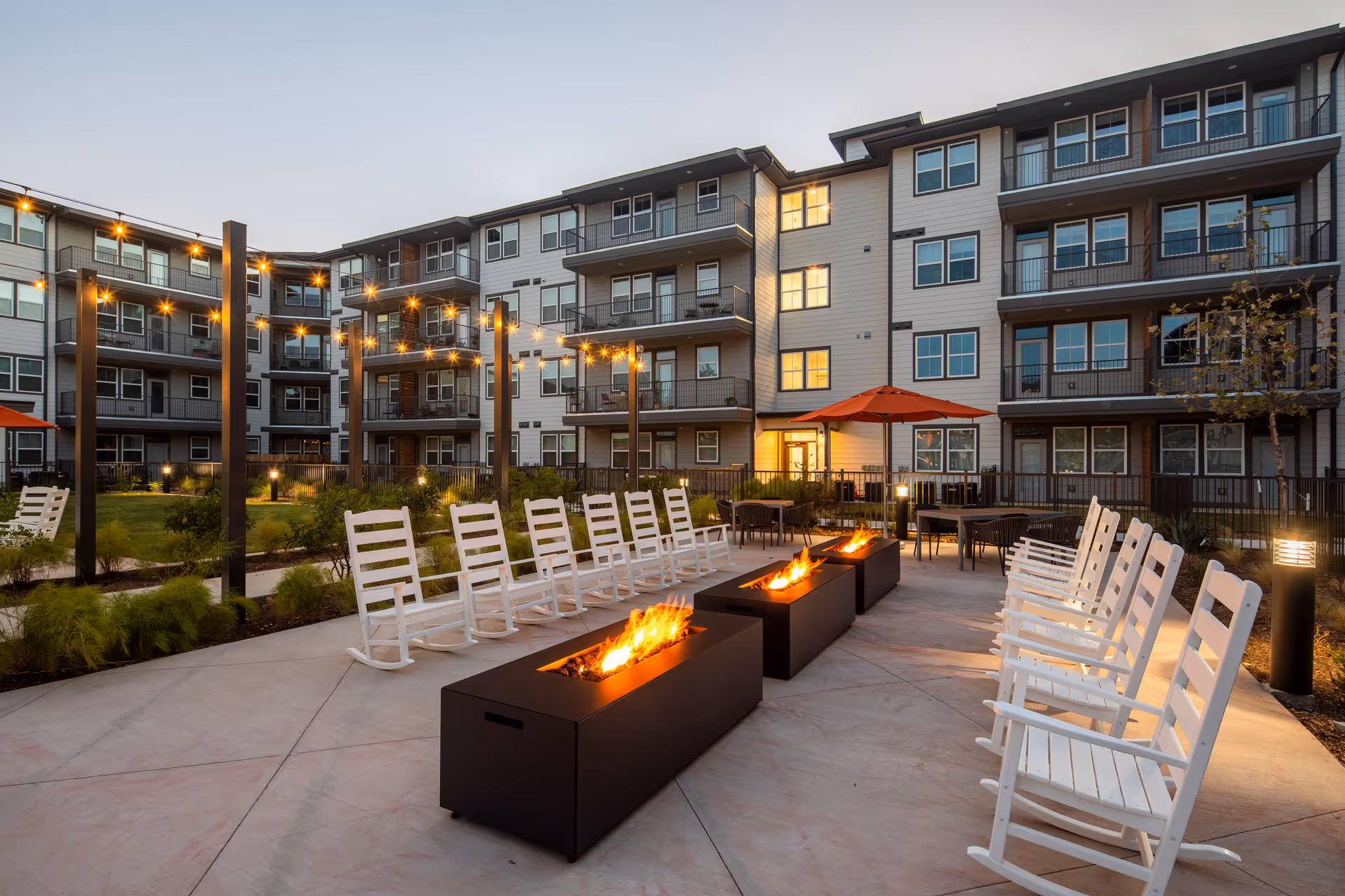 Outdoor courtyard area of a senior living facility with multiple white rocking chairs arranged around three rectangular fire pits. String lights are hung above the seating area, and there are tables with orange umbrellas in the background. The multi-story building with balconies surrounds the courtyard.