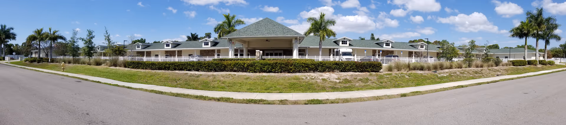 Panoramic view of a single-story senior living building with a covered central entrance, wraparound porch, palm trees, and a road in front under a blue sky.