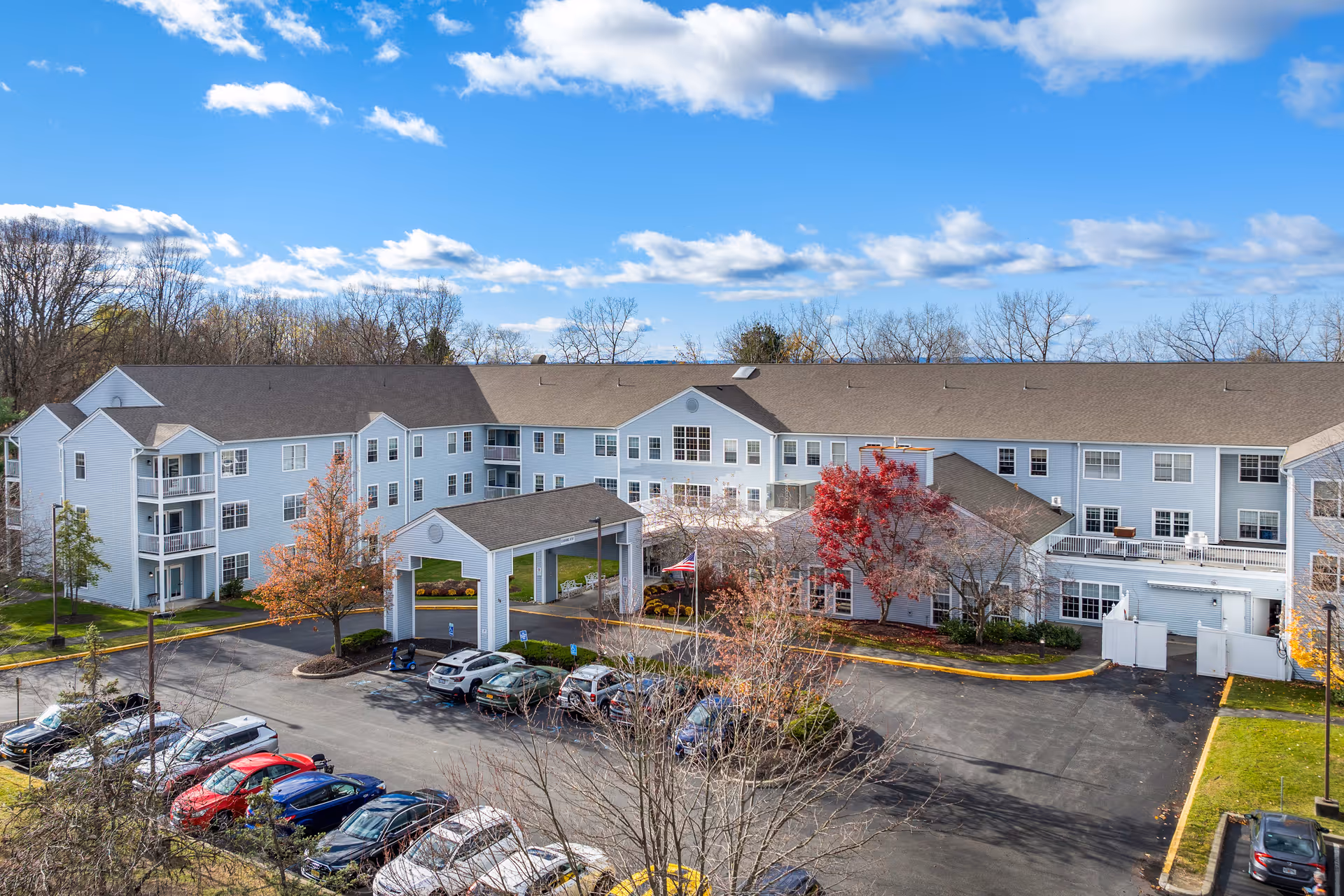 Exterior view of a large three-story senior living community building with a covered entrance, multiple windows, and a parking lot with several cars in front. Trees with autumn foliage surround the building under a partly cloudy blue sky.