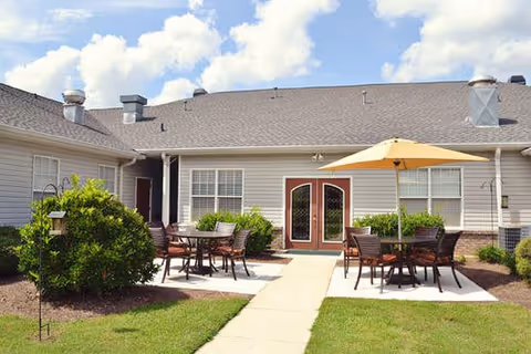 Outdoor patio area at Brookside Commerce with multiple tables and chairs, one table shaded by a large yellow umbrella, surrounded by green bushes and grass under a partly cloudy sky.