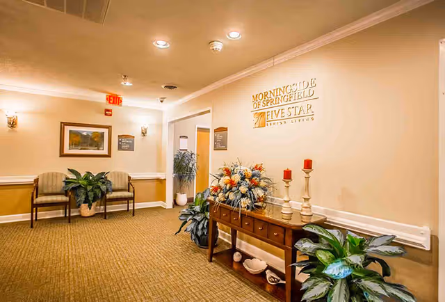 Reception hallway with a wooden console table topped with flowers and candles, potted plants, chairs, and a wall sign reading 'Morningside of Springfield'.