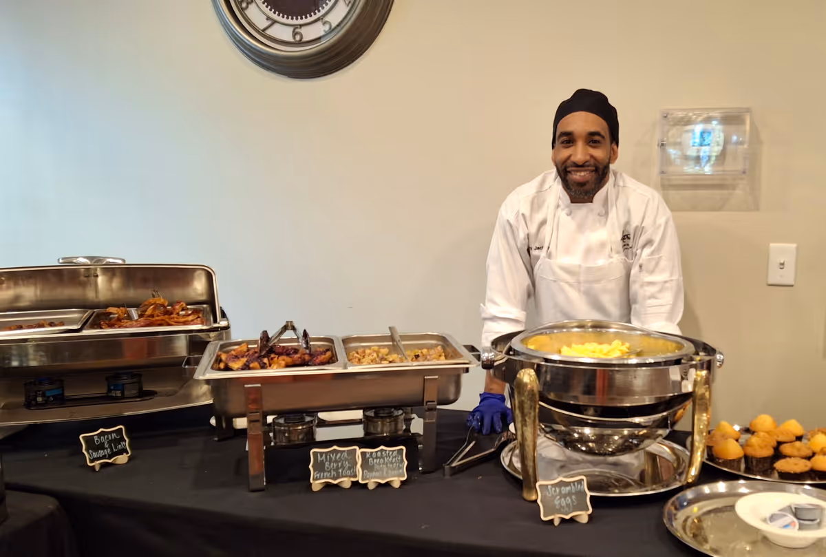 A chef wearing a white uniform and black head covering stands behind a buffet table with various hot food trays including bacon sausage links, mixed berry french toast, roasted breakfast potatoes, scrambled eggs, and muffins. The setting appears to be indoors with a clock on the wall behind him.