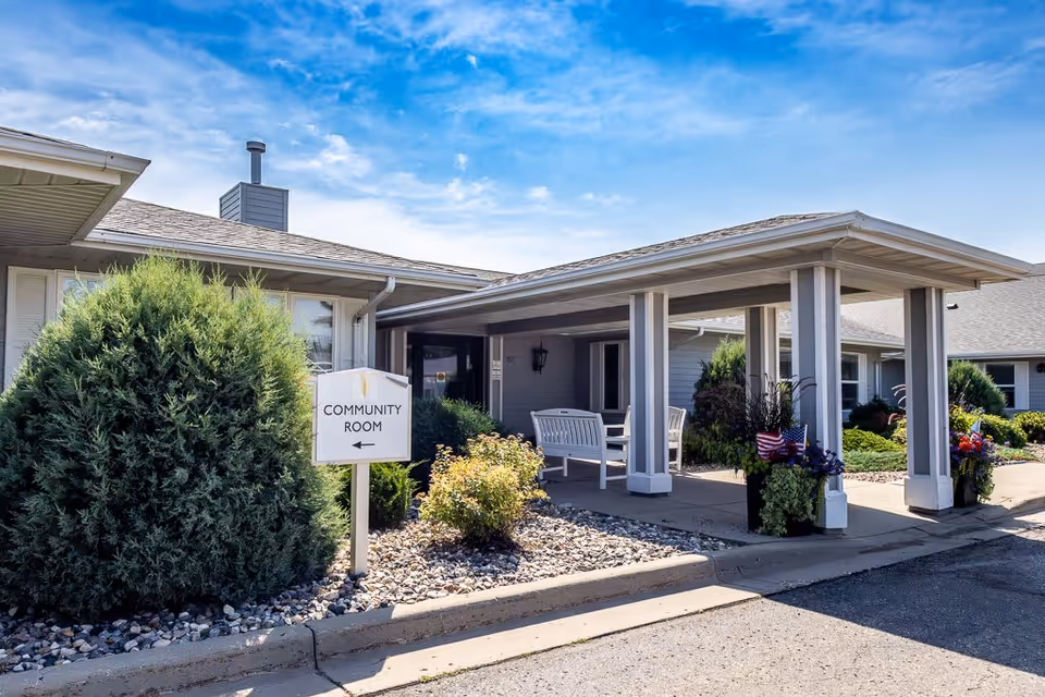 Exterior view of a single-story assisted living facility building with a covered entrance area featuring white benches and potted plants with flowers and small American flags. A sign near the entrance points to the community room. The sky is clear with some clouds.