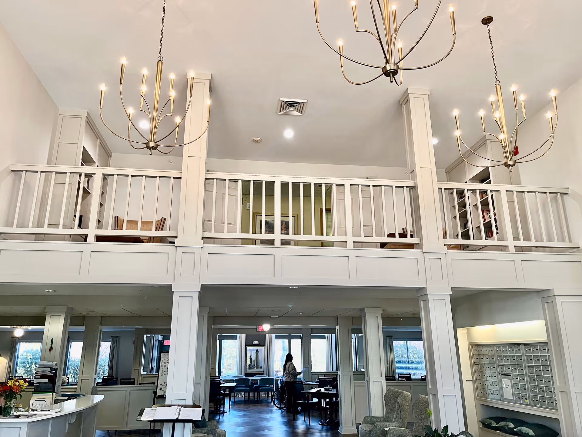 Interior view of a senior living facility with a high ceiling and two large chandeliers. The space features a white railing balcony on the upper level, several columns, a reception desk on the left, seating areas with chairs, and a wall of mailboxes on the right. A person is standing in the background near tables and chairs.