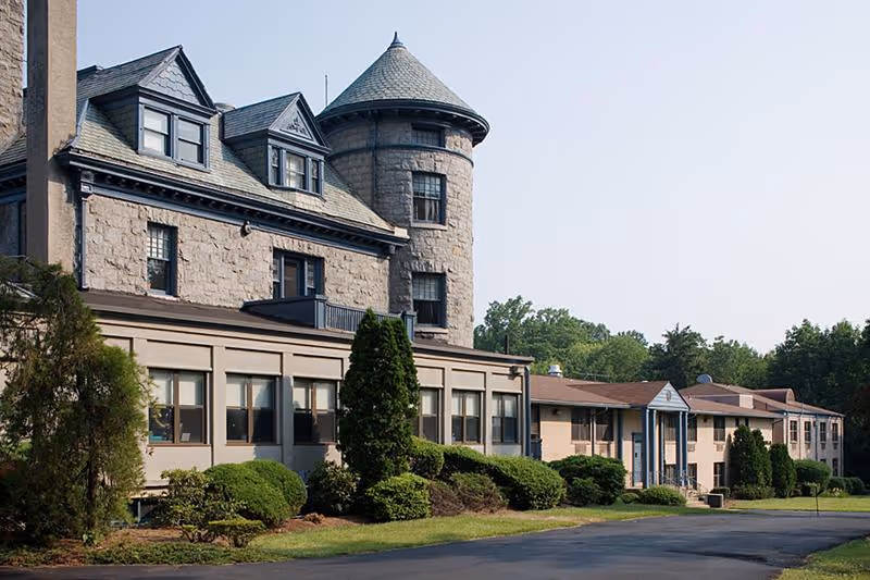 Exterior view of a large stone and brick building with a turret and multiple windows, surrounded by trimmed bushes and trees under a clear sky.