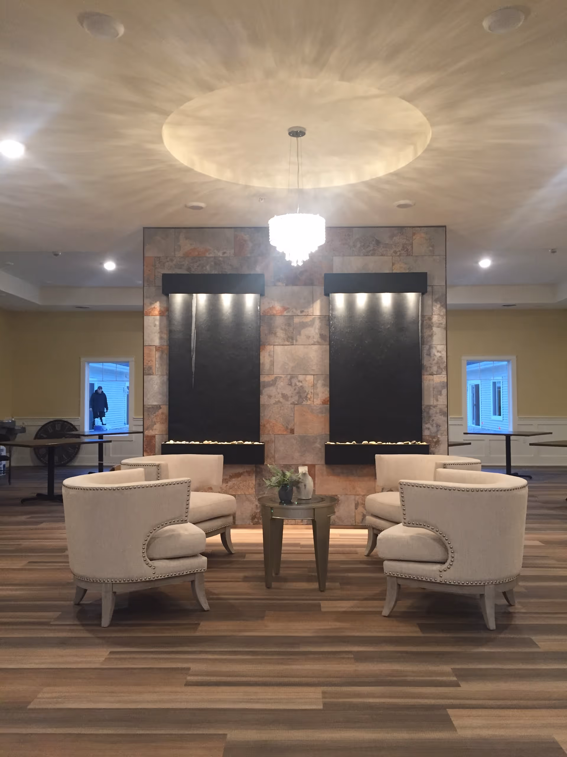 A seating area with four beige upholstered armchairs arranged around a small round wooden table with a plant and decorative items. Behind the seating area is a wall with two vertical water features and a hanging chandelier above, casting light patterns on the ceiling. The room has wood-patterned flooring and windows on either side of the water feature wall.